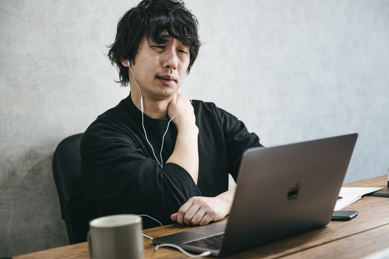 ノートPCで作業しながら、つらそうに首を押さえる男性（イメージ写真）